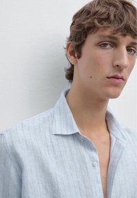 Una giovane persona con capelli corti e ondulati indossa una camicia a righe blu chiaro. Di fronte alla telecamera, contro un muro bianco semplice, mostra un'espressione seria.