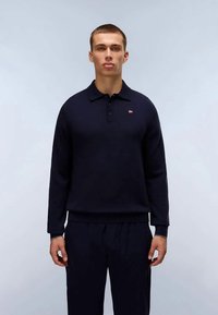 Tamsiai mėlynas megztinis ilgomis rankovėmis ir apykakle, su mažuoju logotipu. Minkšta tekstūra ir laisvas kirpimas, derinamas su atitinkančiomis kelnėmis.