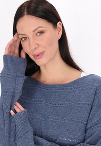 Kék kötött pulóver bordázott mandzsettákkal és nyitott szemekkel készült mintákkal. Széles nyakkivágással és puha textúrával rendelkezik, laza szabású.