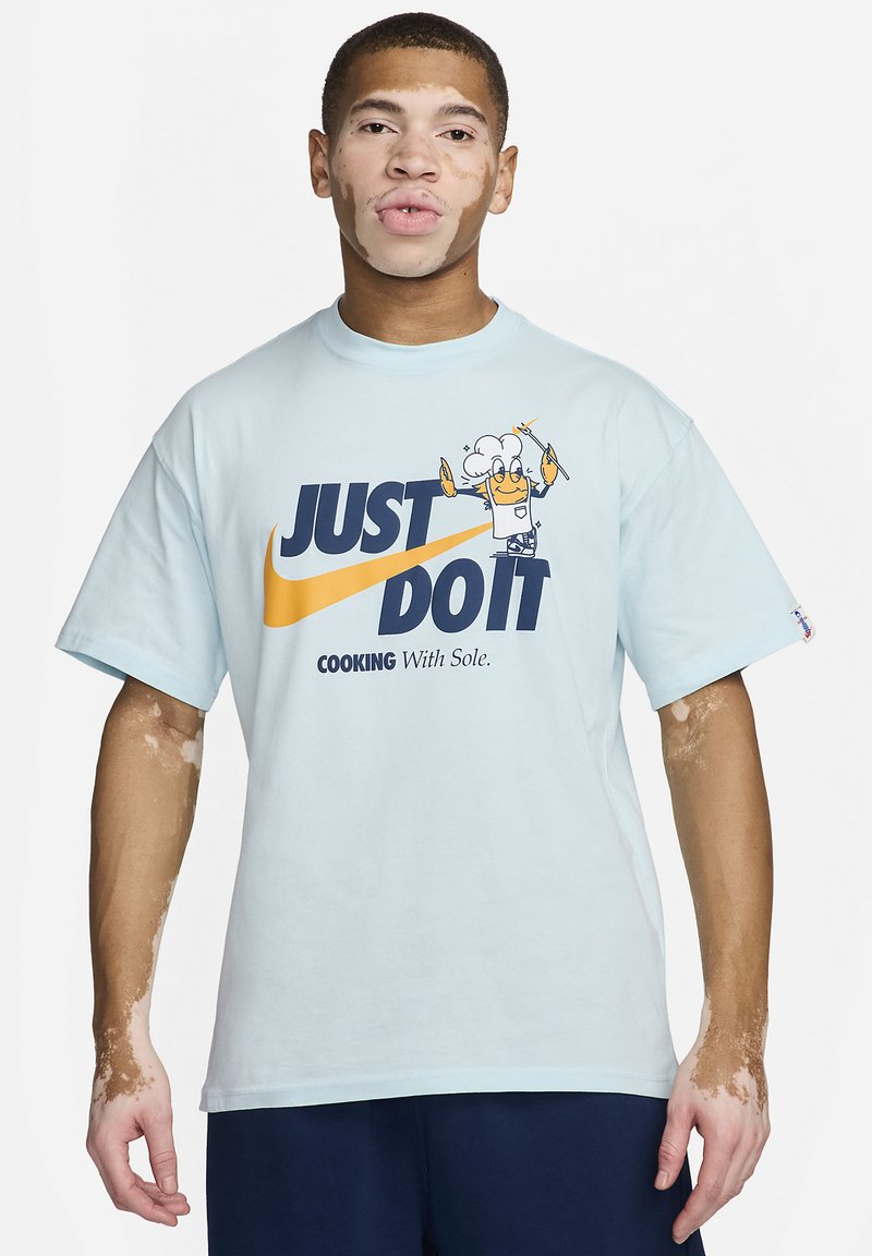 T-shirt en coton bleu clair avec un grand graphisme portant "JUST DO IT," un chef en dessin animé, et "COOKING With Sole" en dessous en bleu foncé. Manches courtes.