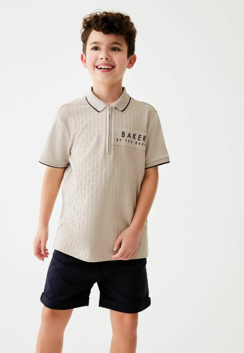 Polo in beige con motivo testurizzato, rifiniture nere e logo, abbinato a pantaloni corti blu navy. Il modello mostra un'espressione sorridente.