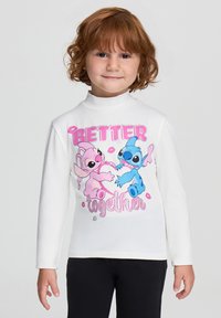 Dziecko z kręconymi, rudymi włosami, ubrane w długi biały top z długim rękawem, na którym znajdują się różowe i niebieskie postacie z kreskówek trzymające serce oraz napis "Lepiej razem".