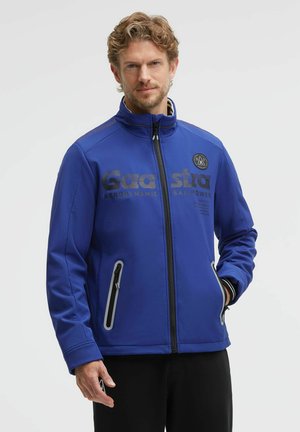 Man with curly hair wearing a blue zip-up jacket with black text and zippered pockets, standing against a plain light background.