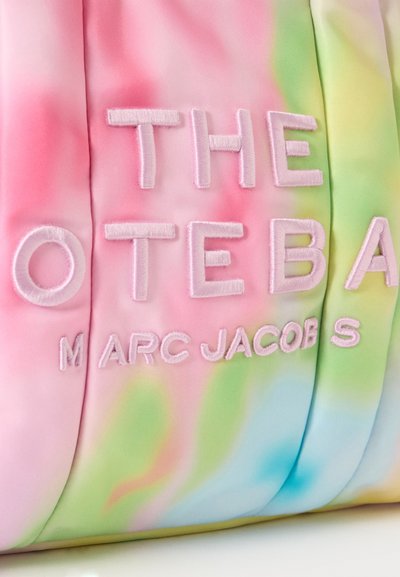 Gros plan d’un sac en tissu tie and dye pastel avec un grand texte brodé indiquant "THE TOTE BAG" et "MARC JACOBS" en lettres rose pâle.