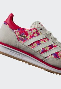 Světlé šedé tenisky Adidas s bílými pruhy, červenou patou a růžovým květinovým vzorem na straně, vyznačují se raženou bílou podrážkou a texturovanými tkaničkami.