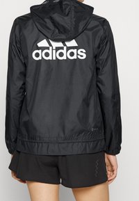 Vista posteriore di una persona che indossa una giacca nera Adidas con cappuccio e un audace logo bianco Adidas, abbinata a pantaloni corti sportivi neri.
