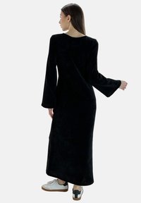 Largo vestido de terciopelo negro con mangas largas, corte holgado, escote redondo y elegante caída, combinado con zapatillas blancas para un contraste.