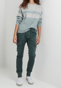 Mujer con un suéter a rayas en tonos azul claro y gris, pantalones jogger cargo en verde oscuro y zapatillas blancas, de pie frente a un fondo liso.