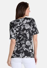 Schwarz gemustertes T-Shirt mit kurzen Ärmeln, das weiße Blumen und Paisleymuster zeigt, aus weichem Stoff, seitlich von hinten betrachtet.