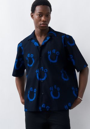 Muž v čiernej krátkorukáve košeli s modrými abstraktnými vzormi usmievajúcich sa tvárí, stojaci na bielom pozadí.