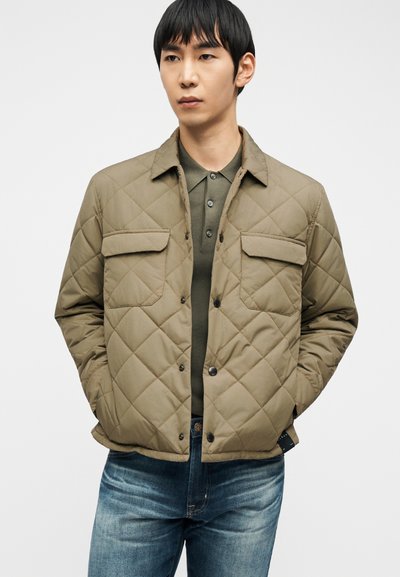 Άνδρας που φοράει μπεζ μπουφάν με καπιτονέ επένδυση πάνω από χακί πράσινη πόλο μπλούζα και μπλε τζιν, στέκεται με τα χέρια στις τσέπες του μπουφάν.