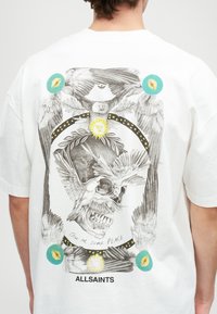 Λευκό βαμβακερό t-shirt με λεπτομερή γραφιστικό σχέδιο που περιλαμβάνει ένα κρανίο, πουλιά, ήλιο και αφηρημένα σχήματα σε μαύρο, κίτρινο και μπλε χρώμα.