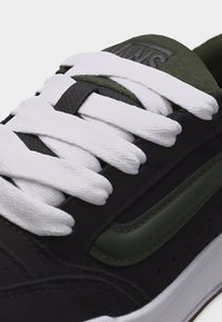 Gros plan sur une basket noire et olive avec des lacets blancs et le logo Vans sur la languette.