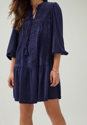 Femme portant une robe bleu marine à manches longues avec une broderie texturée, un décolleté à liens avec pampilles et une partie jupe froncée.