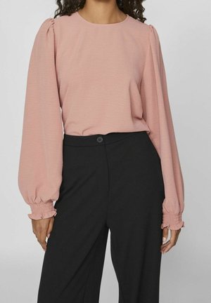 Frau trägt eine langärmelige rosa Bluse mit gerafften Bündchen und schwarze, hochgeschneiderte Hose, steht vor einem einfarbigen Hintergrund.