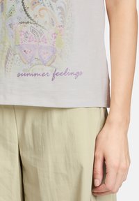 Lichtgrijze t-shirt met daarop een vlinderafbeelding in pastelkleuren, met daaronder sprankelende tekst "zomergevoelens", gecombineerd met beige broek.