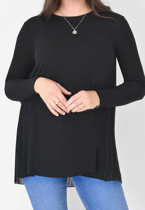 Czarna bluzka z długim rękawem o zwiewnym kroju i bocznych rozcięciach, zestawiona z niebieskimi dżinsami. Złota biżuteria na dłoniach.