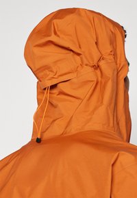 Veste imperméable orange avec une surface texturée et une capuche ajustée, dotée de cordons de serrage pour un ajustement personnalisable.