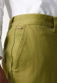 Pantalon en coton vert olive avec une texture lisse, agrémenté d'une étiquette rose et de poches latérales. La ceinture est cousue avec un fil assorti.
