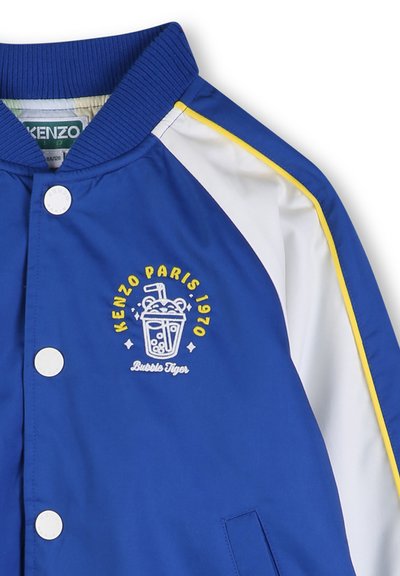 Blauwe nylonjas met witte mouwen en gele accenten. Voorkantlogo toont een beker met een rietje en bubbels. Drukknoopsluiting.