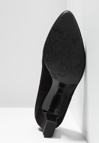 Semelle de chaussure noire à talon haut et bout pointu montrant des motifs décoratifs gaufrés en forme de pièces de monnaie et le nom de la marque "Peter Kaiser" en taille 4½.