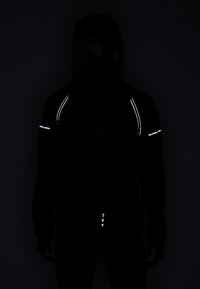 Personne portant une veste noire avec des bandes réfléchissantes sur les épaules, le haut des bras et le bas du dos, debout dos à un arrière-plan sombre.