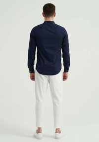 Chemise à manches longues marine avec col montant, associée à un pantalon texturé blanc et des baskets blanches, vue de dos.