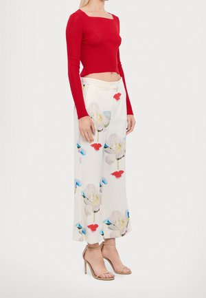 Mujer con top de manga larga rojo recortado, pantalones anchos blancos con gran estampado floral y sandalias de tacón beige, de pie de perfil.