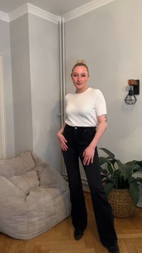 Bela majica s kratkimi rokavi, združena z visokim pasom črnih razširjenih kavbojk. V ozadju je teksturirana svetlo siva kavč in zeleno rastlino.