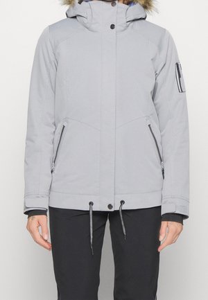 Personne portant une veste d'hiver à capuche gris clair avec bordure en fourrure, fermeture éclair avant avec boutons-pression, poches zippées, et pantalon noir.