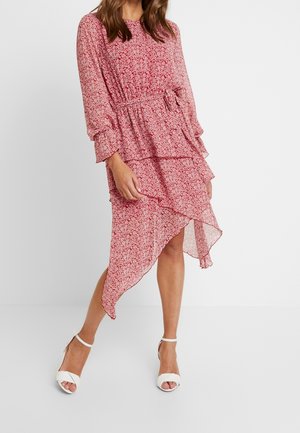 Kvinde iført en lyserød blomsterprintet langærmet kjole med asymmetrisk skørt og hvide hælsandaler, der står mod en ensfarvet baggrund.