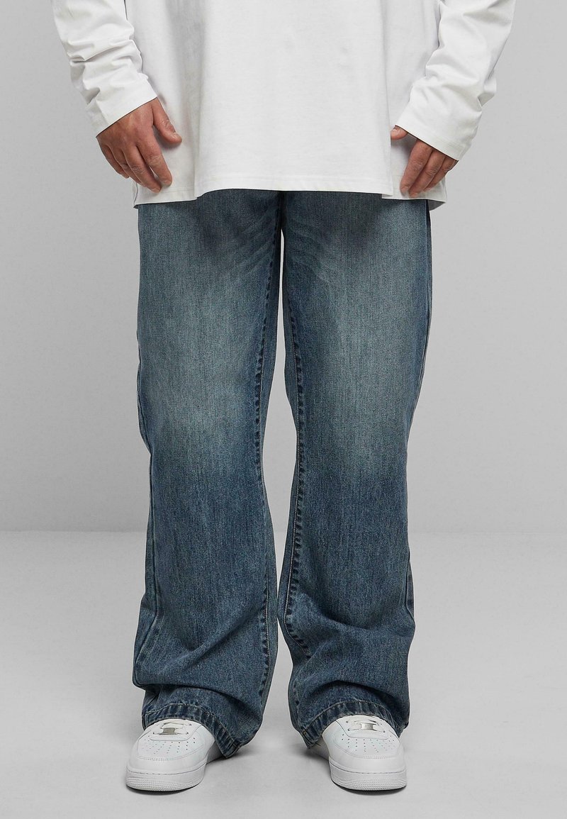 Baggy blue denim jeans with a faded wash, paired with a long white shirt and white sneakers. The jeans have a straight cut and visible stitching.