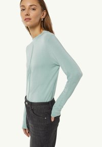 Mintgrünes Longsleeve-Oberteil mit rundem Halsausschnitt und einer mittigen Naht. Kombiniert mit dunkler Denim-Jeans, die eine vordere Tasche und Gürtelschlaufen aufweist.