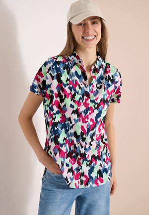 Lächelnde Frau trägt eine beige Kappe mit dem Schriftzug "OPTIMISMUS", ein buntes abstraktes kurzärmeliges Hemd zum Knöpfen und blaue Jeans mit einer Hand in der Tasche.