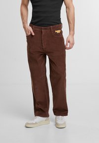 Brown corduroy pants with a straight leg design. Features textured fabric and a logo patch on the waistband. Worn with white sneakers.