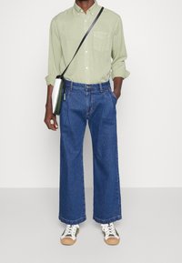 Camicia verde chiaro a maniche lunghe, jeans larghi blu, sneakers bianche con suola beige e una piccola borsa a tracolla verde.