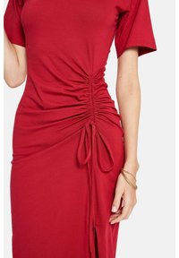 Rotes Kleid aus weichem Stoff, mit kurzen Ärmeln, seitlicher Raffung und einer gebundenen Detail. Das Design beinhaltet einen schlitz bis zur Oberschenkelhöhe.