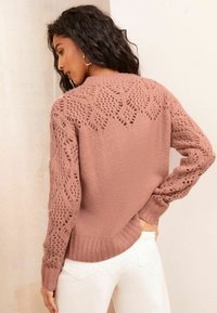 Rosa gestrickter Pullover mit offenen Spitzenmuster an Schultern und Ärmeln, mit gerippten Bündchen und Saum, kombiniert mit weißen Hosen.