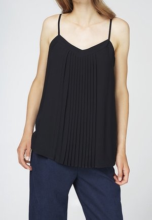 Vrouw die een zwart mouwloos geplooid topje met dunne bandjes en donkerblauwe broek met wijde pijpen draagt, staand tegen een effen achtergrond.