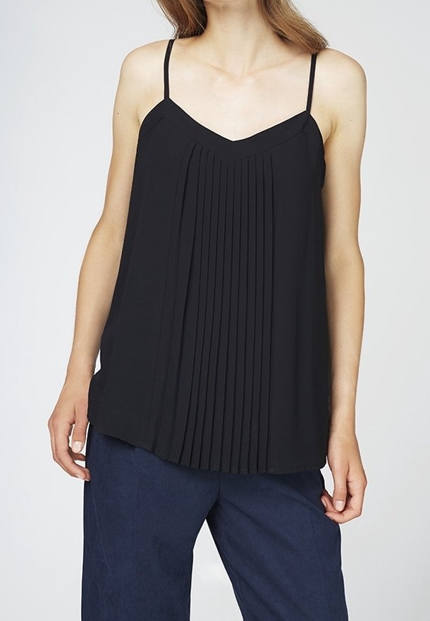 Kobieta ubrana w czarny plisowany top na cienkich ramiączkach bez rękawów oraz ciemnoniebieskie szerokie spodnie, stojąca na jednolitym tle.