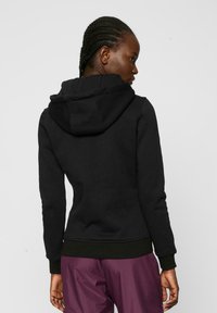 Felpa nera con un fit aderente, tasca kangaroo frontale e cappuccio con cordoncino. Polsini e vita a costine. Texture morbida, branding minimale.