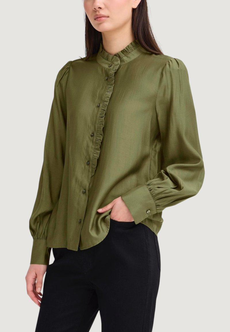 Blusa verde oliva con colletto a volant, chiusura frontale con bottoni e maniche lunghe a sbuffo. Realizzata in tessuto liscio. Abbinata a pantaloni neri.