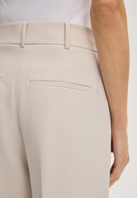 Primo piano della vita posteriore e dei dettagli della tasca su pantaloni beige su misura indossati con una maglietta bianca, con il braccio destro rilassato lungo il corpo.