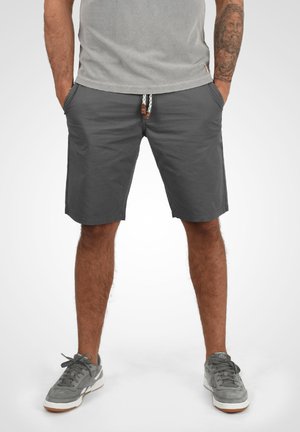 Homme portant un short gris foncé avec cordon, des baskets grises, les mains dans les poches, et une chemise gris clair, debout devant un fond blanc.