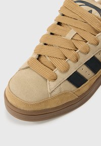 Artimas smėlio spalvos zomšiniai kedai su storiomis rusvomis raištelėmis, juodomis šoninėmis juostelėmis ir ruda guminė pado dalimi ant baltos dėmės.