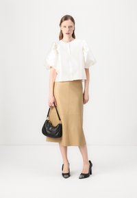 White satin puff-sleeve top paired with a beige knee-length skirt. Model holds a black handbag and wears black heels with metallic toes.