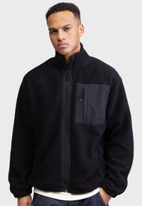 Homme portant une veste en polaire noire avec une fermeture éclair à l'avant et une poche poitrine zippée, se tenant contre un fond gris clair.