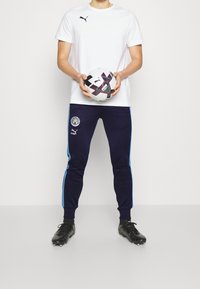 Weißes Kurzarmhemd mit schwarzem Logo, marineblaue Jogginghose mit hellblauen Seiten, hält einen gemusterten schwarz-weißen Fußball und trägt schwarze Fußballschuhe.