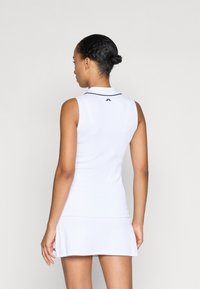 Bílá bezrukávová polo šaty s ribovanou texturou, černými límečkovými akcenty a logem na zadní straně, s přiléhavým střihem a rozšířenou sukní.