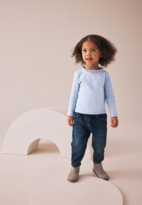 Hellblaues Fleecepullover mit langen Ärmeln und gerafftem Ausschnitt, kombiniert mit dunkelblauen Jeans und grauen Knöchelstiefeln. Stehend vor einem minimalistischen Hintergrund.